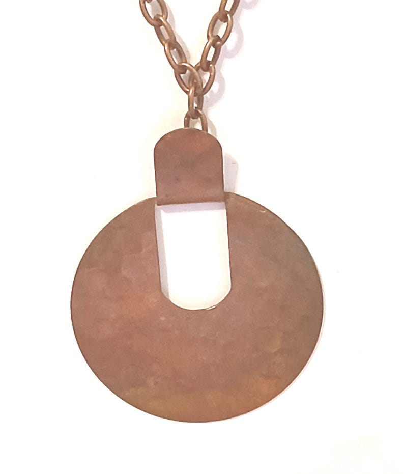 May include: A copper-coloured pendant necklace. The pendant features a circular design with a rectangular top and a U-shaped cutout. The chain is made of matching copper-coloured links. The necklace is set against a white background.