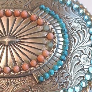 May include: Close-up of a silver-toned belt buckle with intricate floral and geometric engravings. The buckle is adorned with turquoise and coral-colored beads, arranged in decorative patterns around the edges and center.