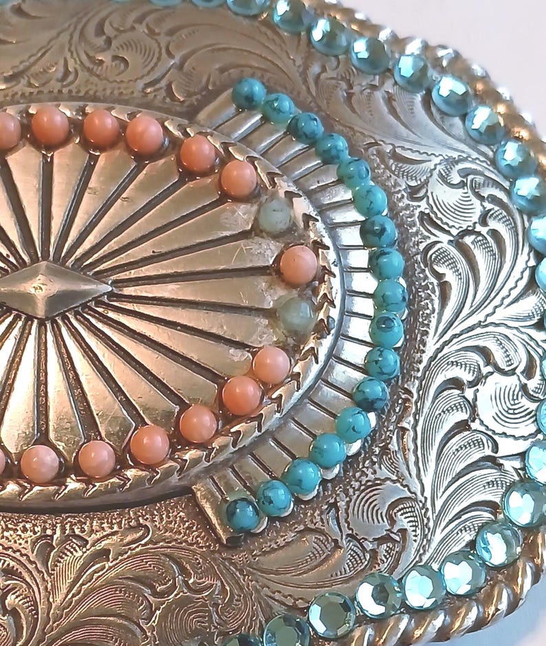 May include: Close-up of a silver-toned belt buckle with intricate floral and geometric engravings. The buckle is adorned with turquoise and coral-colored beads, arranged in decorative patterns around the edges and center.