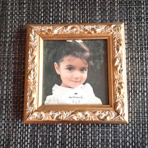 May include: A square picture frame with an ornate, embossed gold finish featuring a decorative scroll pattern. The frame contains a photograph of a child wearing a white top, with a Hallmark 5 x 5 label visible on the lower portion of the image.