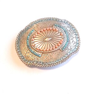 May include: Silver-toned oval belt buckle with intricate floral engravings. The buckle features a central oval design with a sunburst pattern, surrounded by coral-colored and turquoise-colored beads. The edges are adorned with turquoise-colored beads.