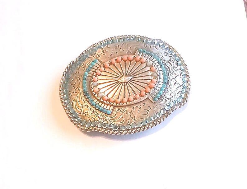 May include: Silver-toned oval belt buckle with intricate floral engravings. The buckle features a central oval design with a sunburst pattern, surrounded by coral-colored and turquoise-colored beads. The edges are adorned with turquoise-colored beads.