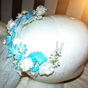 May include: A white headpiece adorned with a floral arrangement of blue and white flowers. The headpiece is decorated with a delicate veil. The flowers are arranged in a circular pattern around the base of the headpiece.