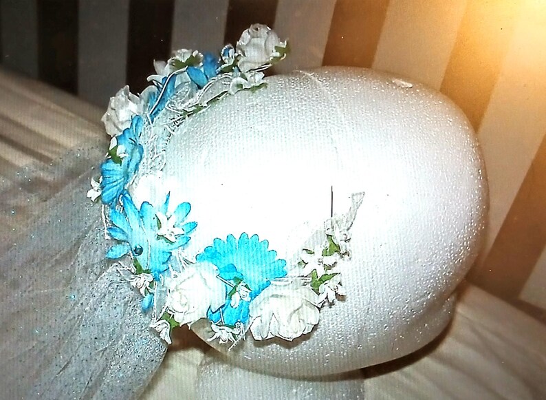 May include: A white headpiece adorned with a floral arrangement of blue and white flowers. The headpiece is decorated with a delicate veil. The flowers are arranged in a circular pattern around the base of the headpiece.
