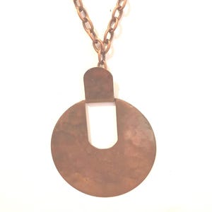 May include: A copper-coloured necklace featuring a large, circular pendant. The pendant has a cutout design and is suspended from a matching chain. The necklace is set against a white background.