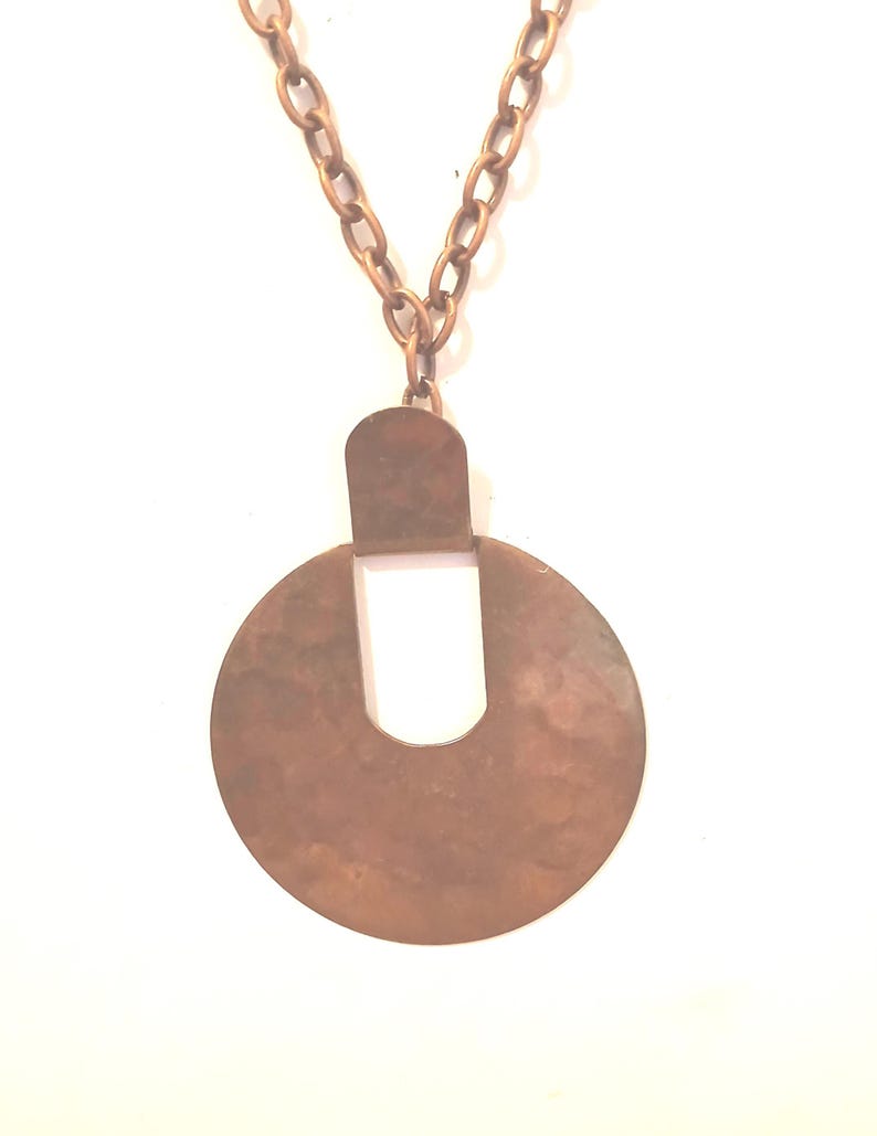 May include: A copper-coloured necklace featuring a large, circular pendant. The pendant has a cutout design and is suspended from a matching chain. The necklace is set against a white background.