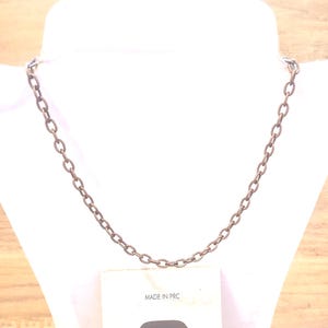 May include: A bronze-coloured chain necklace displayed on a white mannequin. The necklace is made of oval-shaped links. The tag below the necklace reads "MADE IN PRC". The background is a light wood.