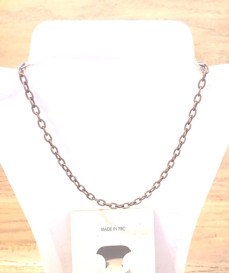 May include: A bronze-coloured chain necklace displayed on a white mannequin. The necklace is made of oval-shaped links. The tag below the necklace reads "MADE IN PRC". The background is a light wood.