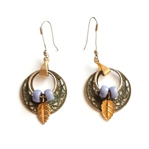 May include: A pair of dangle earrings featuring a gold-toned metal leaf charm suspended from a silver-toned circular frame with a textured lattice pattern. Each earring includes two light blue cylindrical beads and a silver-toned hook wire.