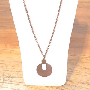 May include: A copper-toned necklace with a circular pendant and a U-shaped cutout. The pendant is suspended from a fine chain, presented on a white stand against a wooden backdrop. The necklace has a vintage or bohemian style.