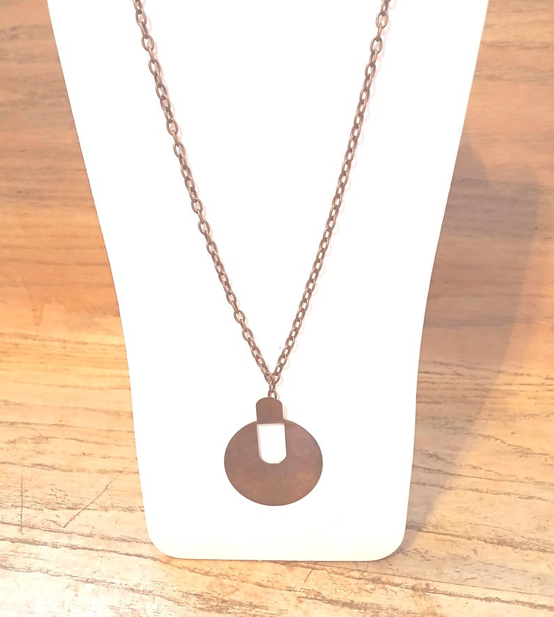 May include: A copper-toned necklace with a circular pendant and a U-shaped cutout. The pendant is suspended from a fine chain, presented on a white stand against a wooden backdrop. The necklace has a vintage or bohemian style.