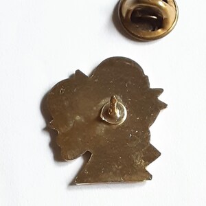 Puede incluir: Pin de metal dorado con el perfil de una cabeza. El pin tiene un cierre de seguridad en la parte posterior.