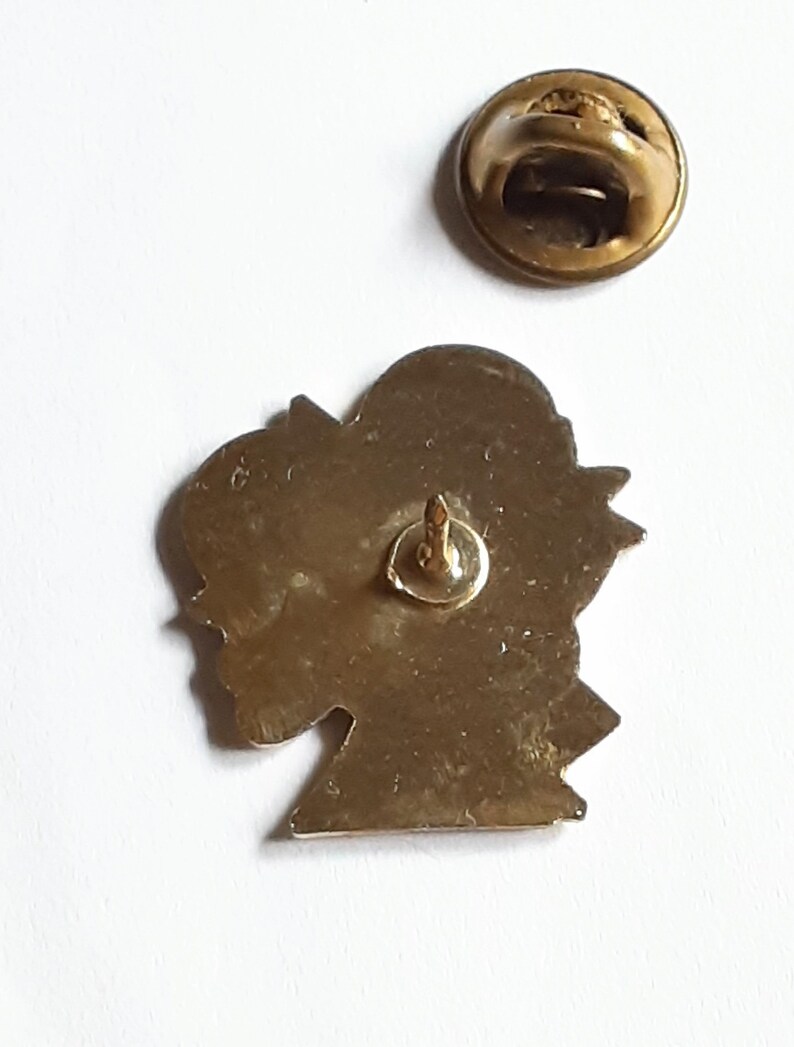 Puede incluir: Pin de metal dorado con el perfil de una cabeza. El pin tiene un cierre de seguridad en la parte posterior.