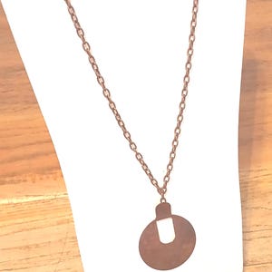 May include: A copper-coloured necklace featuring a circular pendant with a rectangular top. The chain is composed of small, linked circles. The pendant is suspended from the chain.