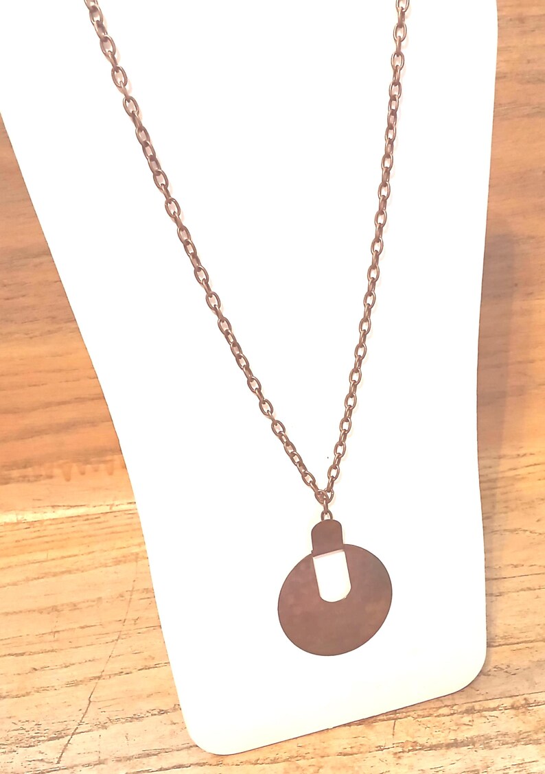 May include: A copper-coloured necklace featuring a circular pendant with a rectangular top. The chain is composed of small, linked circles. The pendant is suspended from the chain.