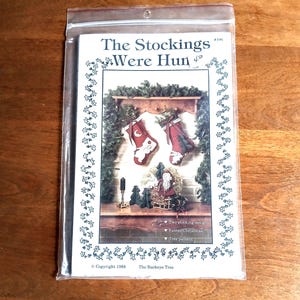 Pode incluir: Um pacote contendo um padrão de artesanato com tema natalino. O padrão apresenta o texto "The Stockings Were Hun" e ilustrações de meias de Natal, Papai Noel e uma árvore de Natal. O pacote é selado em um saco plástico transparente.