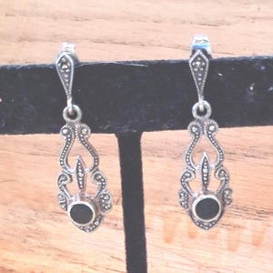 May include: A pair of silver-toned drop earrings with an ornate, filigree design. Each earring features a small, round black stone at the center and is accented with tiny, sparkling details. The earrings are displayed against a wooden background.