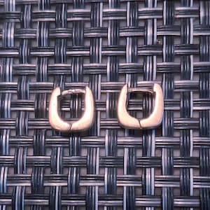 May include: A pair of small, silver-toned hoop earrings with a square-shaped design. Each earring features a hinged closure mechanism, creating a continuous, smooth metallic loop. The earrings have a polished finish and a simple, geometric structure.