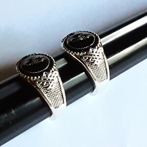 May include: Two silver-toned rings featuring a circular black stone inlay at the centre. The band of each ring displays an intricate, raised metalwork pattern with a textured, cross-hatched design on the sides, creating a detailed appearance.