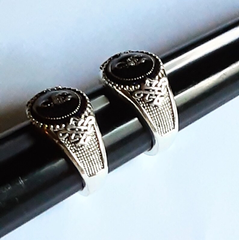May include: Two silver-toned rings featuring a circular black stone inlay at the centre. The band of each ring displays an intricate, raised metalwork pattern with a textured, cross-hatched design on the sides, creating a detailed appearance.