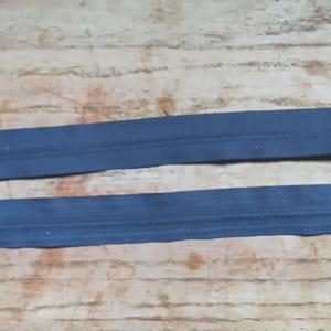 May include: Two navy blue zippers with white tags attached, lying parallel on a wooden surface. The zippers have metal pulls and teeth. The tags have a barcode and text.