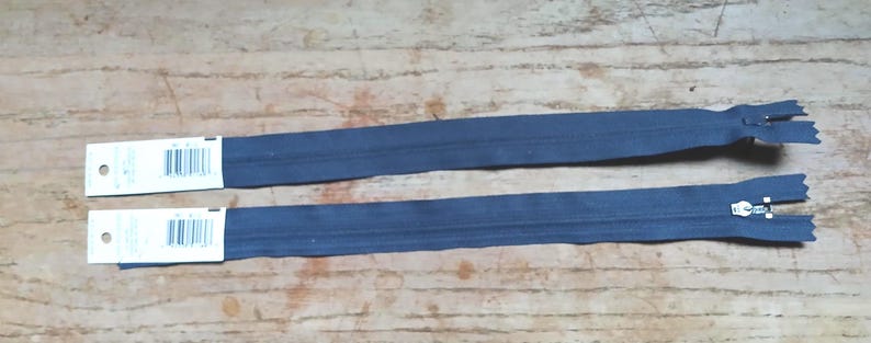May include: Two navy blue zippers with white tags attached, lying parallel on a wooden surface. The zippers have metal pulls and teeth. The tags have a barcode and text.