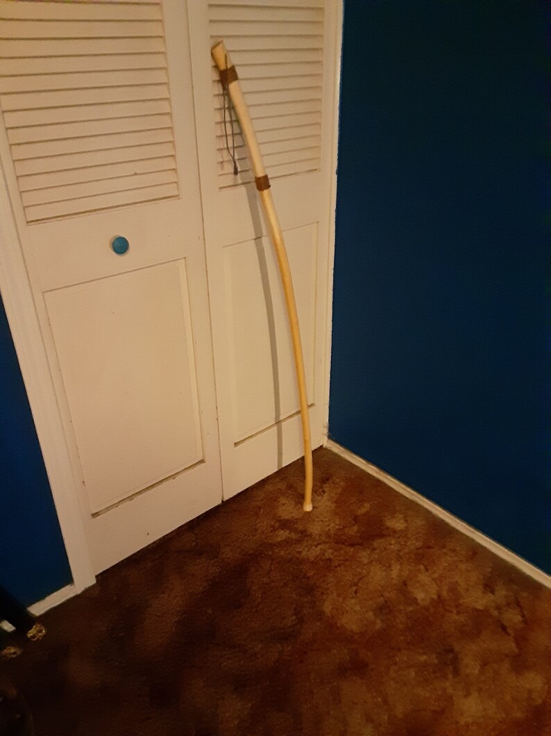Op de afbeelding: Een lange, lichtbruine wandelstok met een donkerbruine leren wikkeling aan de bovenkant. De stok leunt tegen een witte deur met een blauwe muur op de achtergrond.