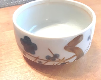 May include: A round ceramic bowl with a white glazed interior and exterior. The exterior features a hand-painted design consisting of dark blue flower shapes, thin brown intersecting lines, and a larger brown brushstroke accent near the rim.