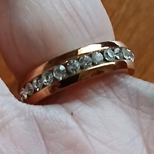 May include: A gold-colored ring with a row of clear, sparkling stones set along the band. The ring is shown on a person's finger, with the focus on the jewelry's design and details. The ring's band has a smooth, polished finish.