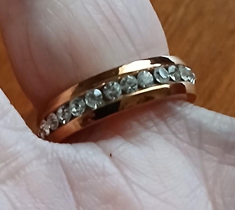 May include: A gold-colored ring with a row of clear, sparkling stones set along the band. The ring is shown on a person's finger, with the focus on the jewelry's design and details. The ring's band has a smooth, polished finish.