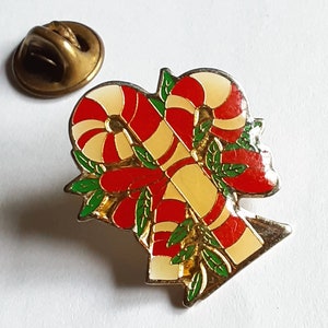 Puede incluir: Un pin de tono dorado con un dise&ntilde;o de bast&oacute;n de caramelo rojo y blanco. Los bastones de caramelo est&aacute;n entrelazados y rodeados de hojas verdes.