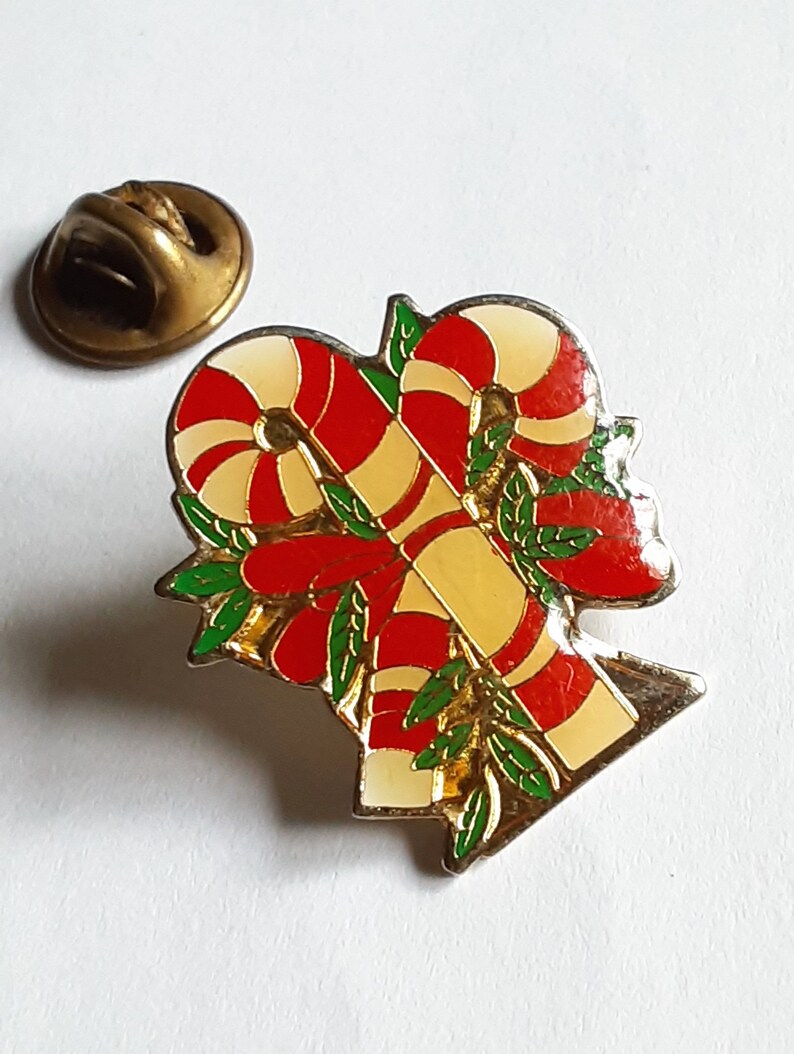 Puede incluir: Un pin de tono dorado con un dise&ntilde;o de bast&oacute;n de caramelo rojo y blanco. Los bastones de caramelo est&aacute;n entrelazados y rodeados de hojas verdes.