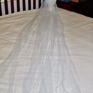 May include: A bridal veil with a floral crown. The veil is long, sheer, and white, with subtle glitter accents. The crown features white and light blue flowers. The veil is displayed on a mannequin head.