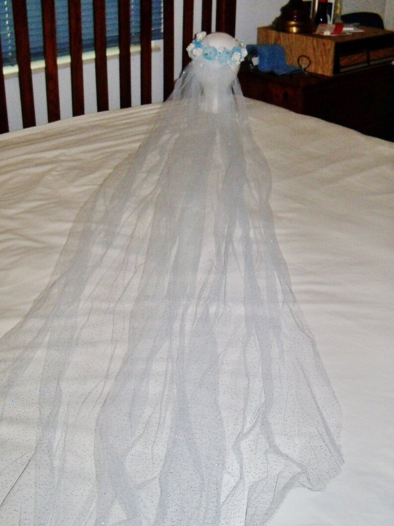 May include: A bridal veil with a floral crown. The veil is long, sheer, and white, with subtle glitter accents. The crown features white and light blue flowers. The veil is displayed on a mannequin head.
