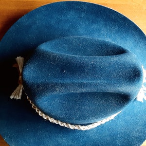 Puede incluir: Un sombrero vaquero de fieltro azul oscuro con una banda trenzada blanca y borlas blancas.