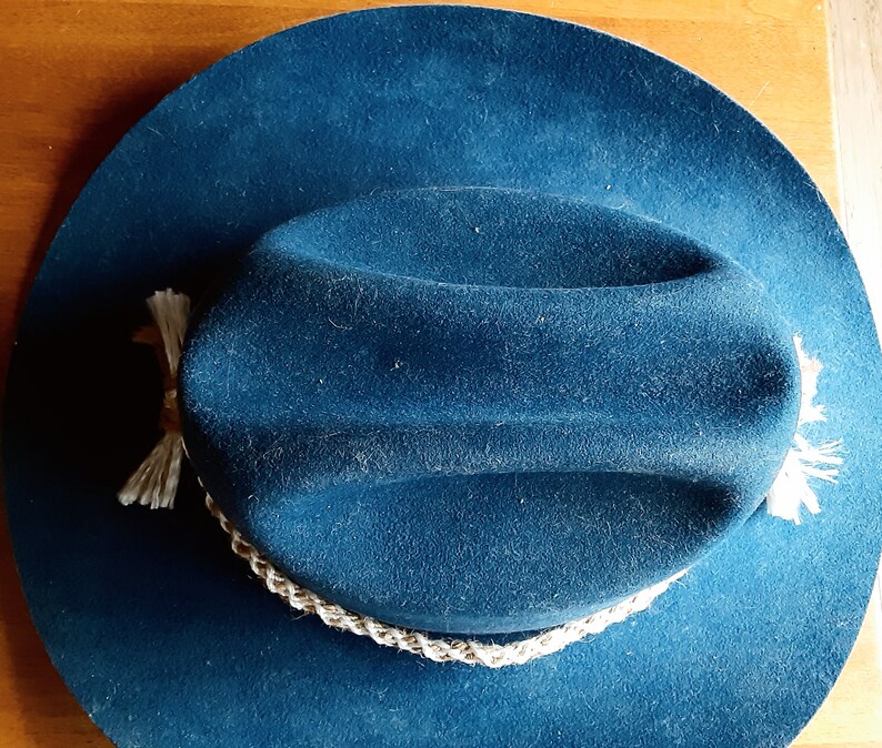 Puede incluir: Un sombrero vaquero de fieltro azul oscuro con una banda trenzada blanca y borlas blancas.