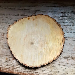 May include: A round wooden slice with a natural wood grain pattern and a rough bark edge. The wood is light brown in color and has a smooth surface.