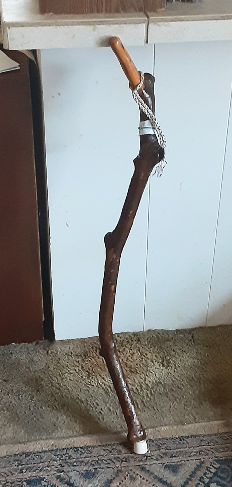 May include: A wooden walking stick with a brown, knobby handle and a white rubber tip. The stick is decorated with a white cord and a green tag.