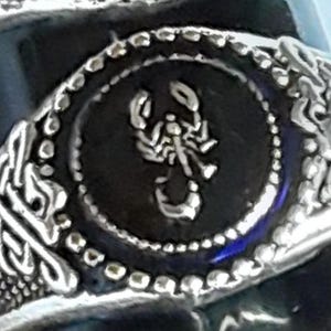 May include: A silver-coloured ring featuring a circular centrepiece with a raised scorpion emblem. The central disc is surrounded by a beaded border, and the band displays intricate, textured knotwork patterns on both sides of the main setting.