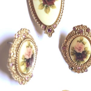 May include: A vintage jewelry set featuring a pendant and matching earrings. The pieces have a gold-tone filigree design and are adorned with oval-shaped cream-colored inserts. Each insert displays a floral design with pink and burgundy roses.