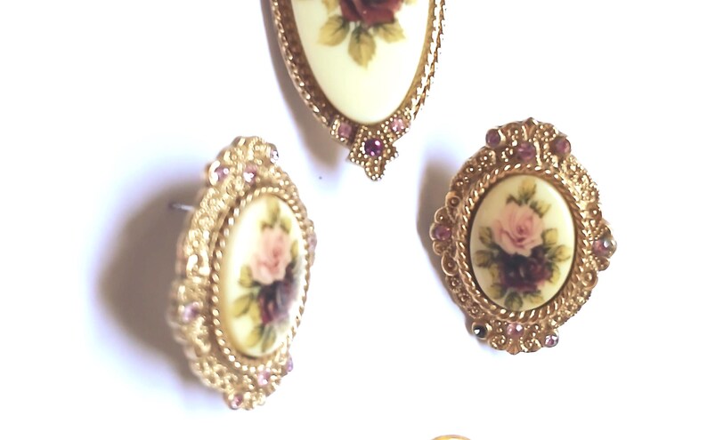 May include: A vintage jewelry set featuring a pendant and matching earrings. The pieces have a gold-tone filigree design and are adorned with oval-shaped cream-colored inserts. Each insert displays a floral design with pink and burgundy roses.