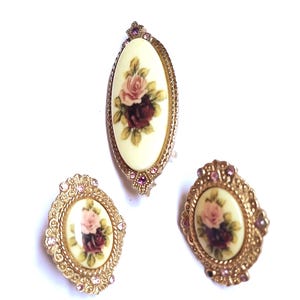 May include: A vintage jewelry set featuring a brooch and two matching earrings. The oval-shaped pieces have a cream-colored background with a floral rose design in pink and burgundy. The gold-toned metal frames are embellished with small pink rhinestones.