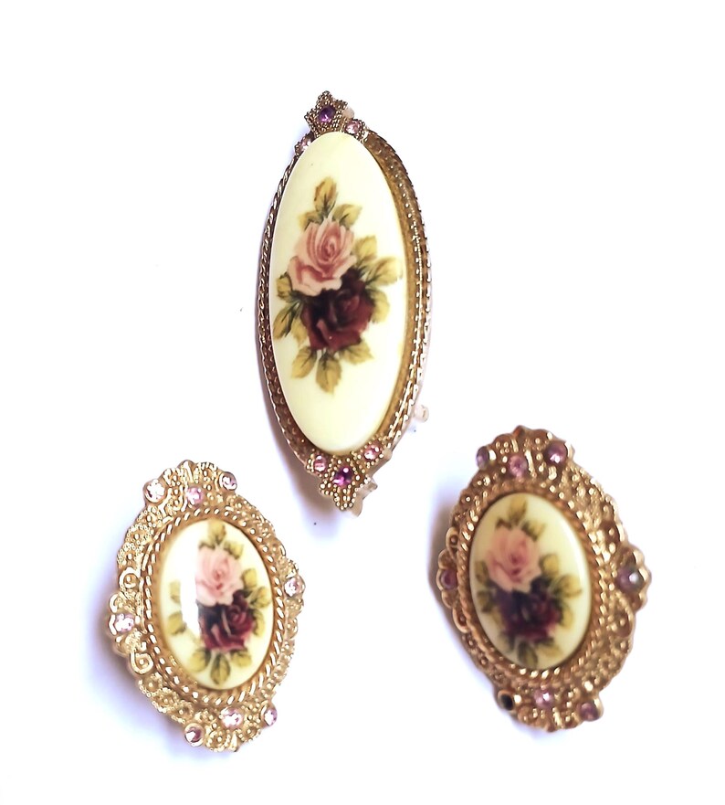 May include: A vintage jewelry set featuring a brooch and two matching earrings. The oval-shaped pieces have a cream-colored background with a floral rose design in pink and burgundy. The gold-toned metal frames are embellished with small pink rhinestones.