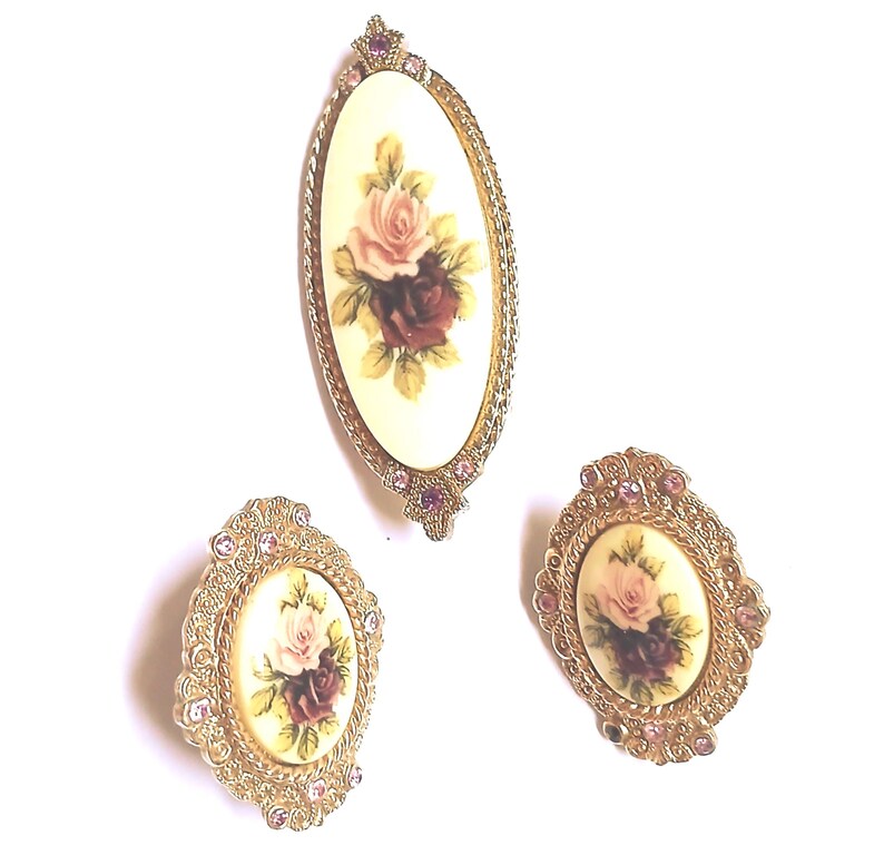 May include: A set of three vintage brooches. Each brooch features an oval cream-colored center with a floral design of pink and burgundy roses. The brooches are framed in gold-tone metal with pink rhinestone accents.