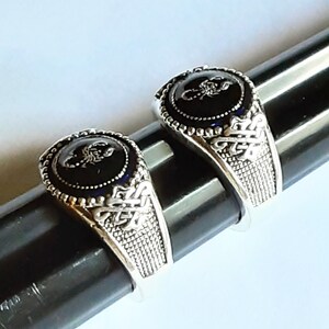 May include: Two silver-toned rings featuring a circular black inlay with a raised silver scorpion emblem. The band has a textured grid pattern on the sides and an intricate knotwork design near the setting, which is bordered by small silver beads.