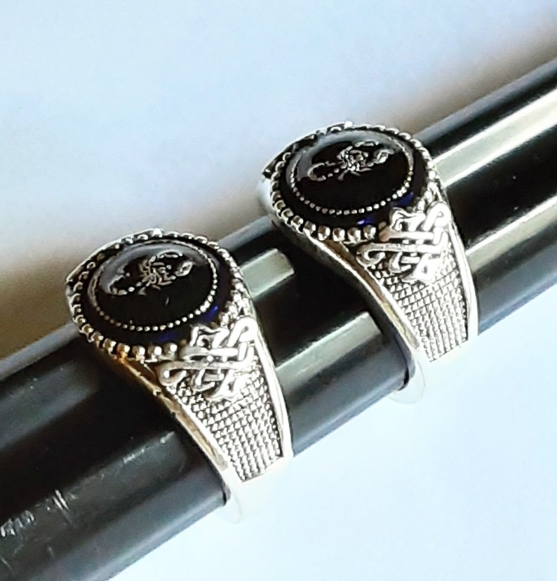 May include: Two silver-toned rings featuring a circular black inlay with a raised silver scorpion emblem. The band has a textured grid pattern on the sides and an intricate knotwork design near the setting, which is bordered by small silver beads.