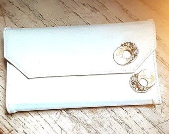 May include: A light-colored clutch with a flap closure. The clutch features two decorative, circular clasps with gold and cream-colored accents. The accessory is made of a soft, possibly leather-like material.