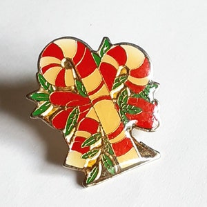 Puede incluir: Un pin de color dorado con un dise&ntilde;o de bast&oacute;n de caramelo rojo y blanco. Los bastones de caramelo est&aacute;n entrelazados y rodeados de hojas verdes.