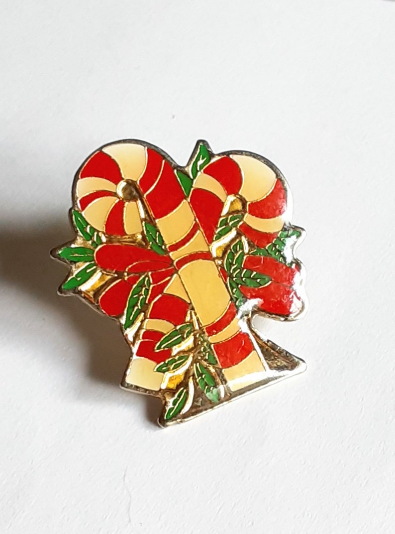 Puede incluir: Un pin de color dorado con un dise&ntilde;o de bast&oacute;n de caramelo rojo y blanco. Los bastones de caramelo est&aacute;n entrelazados y rodeados de hojas verdes.