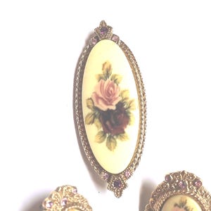 May include: An oval brooch and two earrings with a floral design. The brooch features a cream-colored oval with a painting of pink and burgundy roses. The brooch and earrings have a gold-colored metal frame with pink accents.